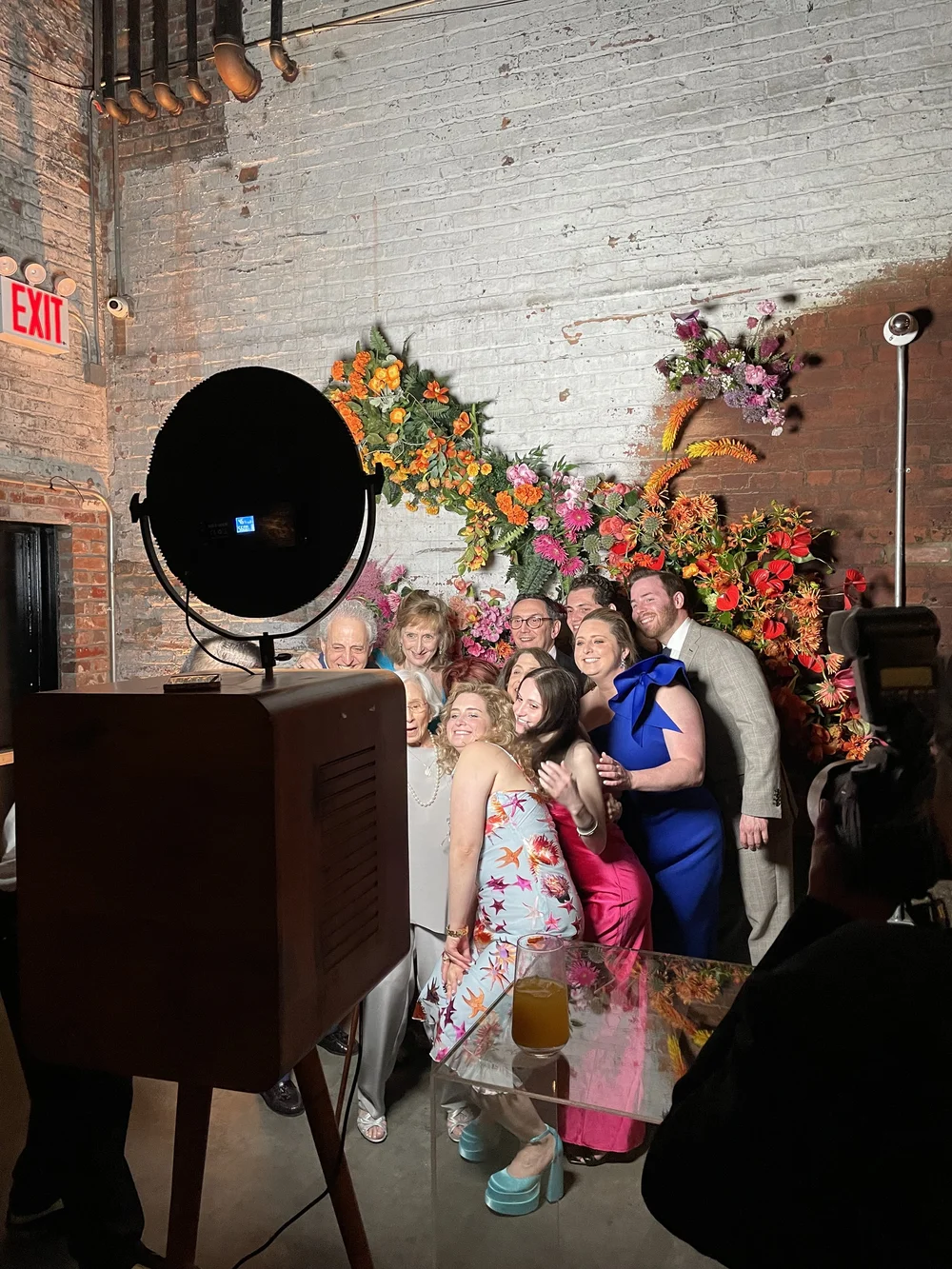Group of people posing for a photo at a party or event with colorful floral wall decoration in the background.