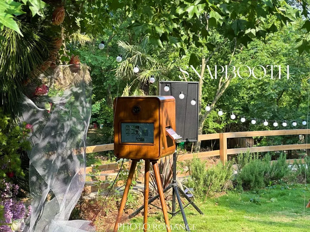 Vintage photo booth with wooden exterior on tripod stand, black speaker behind it, surrounded by greenery, string lights, and floral decorations, in outdoor garden setting.