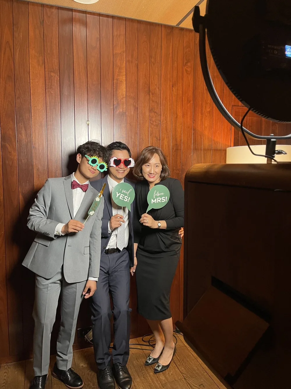 Three people in formal attire posing for a photo, holding signs that say "I said YES!" and "Future MRS." They are wearing humorous sunglasses, and the photo is being taken with a professional camera in a wood-paneled room.