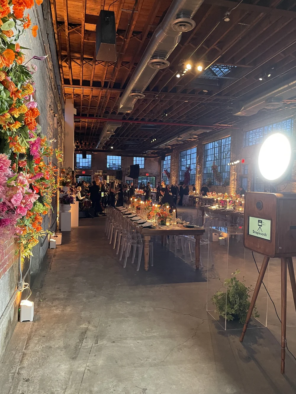 Interior of a decorated event space with long tables set for a dinner, floral centerpieces, and guests in the background. The room features exposed brick walls, large windows, and a high wooden ceiling with industrial ductwork and lighting.
