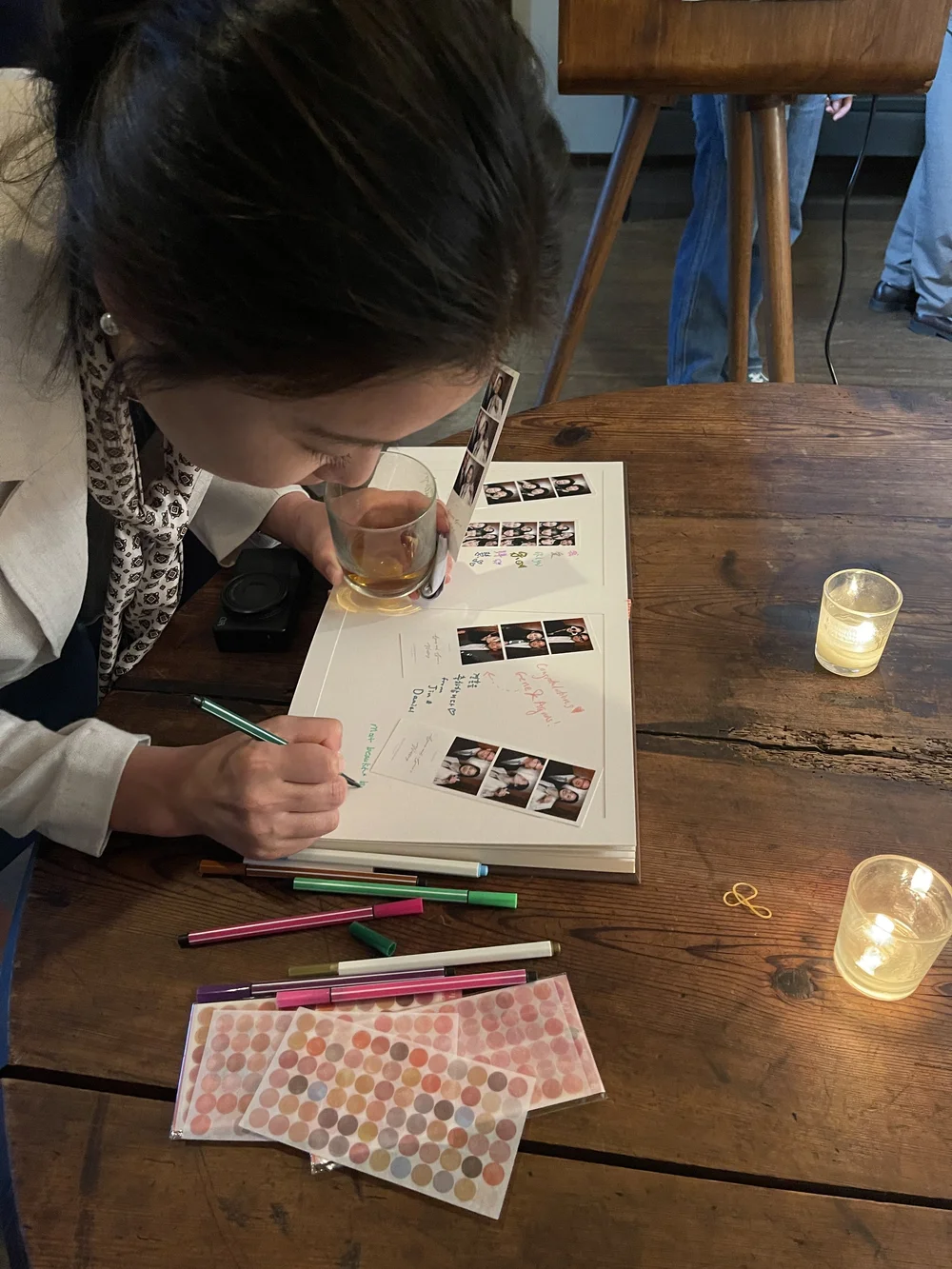 A woman writes in a scrapbook with colorful pens on a wooden table, surrounded by candles, photo booth pictures, and supplies.