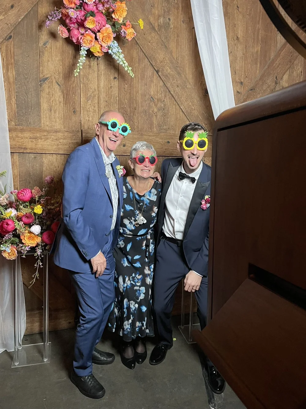Three people at a joyous event wearing colorful pineapple-shaped sunglasses, standing in front of a wooden wall adorned with pink and multicolored flowers.