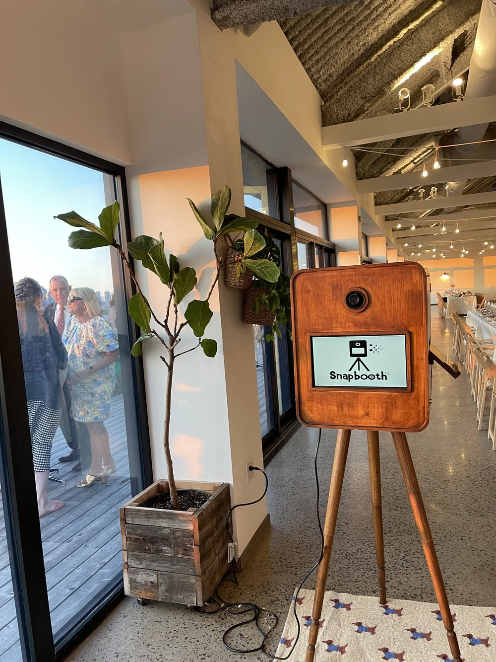 A photo booth station with a wooden box and a screen displaying the Snapbooth logo, set up inside a venue with large windows, potted plants, and people talking outside.