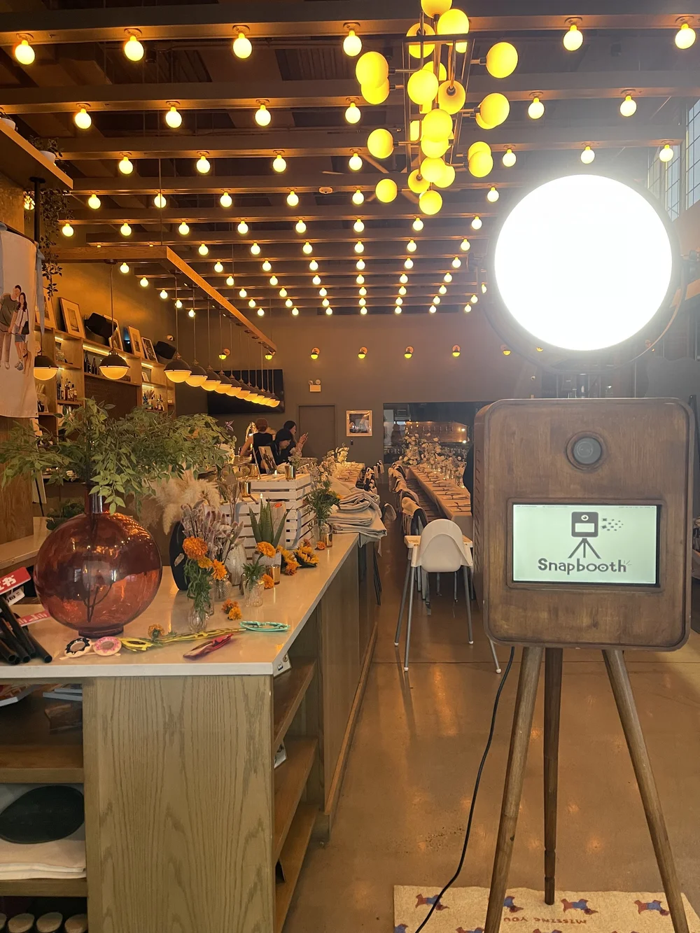 Inside a warmly lit event space or restaurant with an inviting atmosphere, decorated with yellow string lights hanging from the ceiling. The space features a long table set for a gathering, adorned with flowers, plants, and decorative items. There is